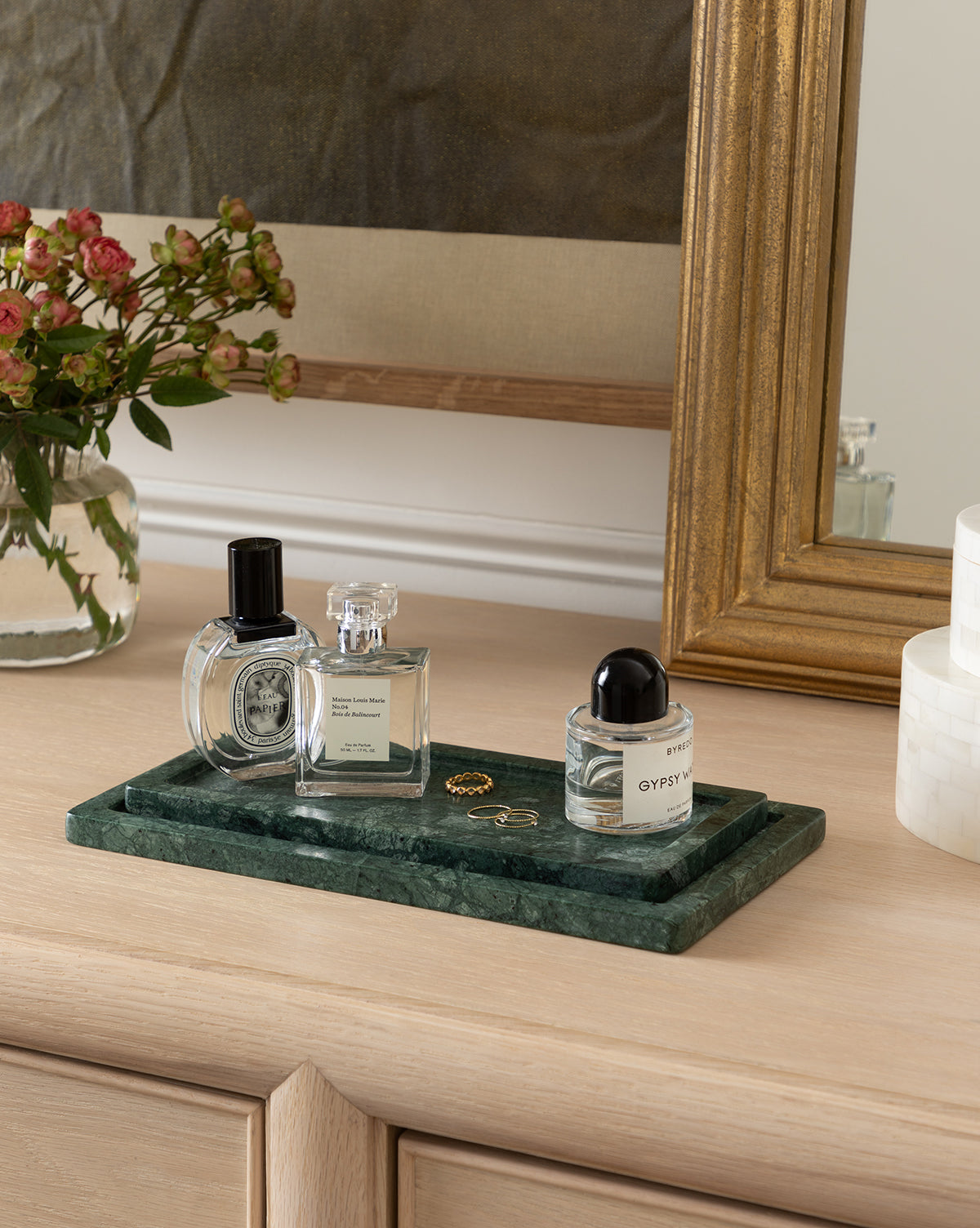 The Green Marble Nesting Tray displays three perfume bottles and two rings on a light wood dresser, styled with a vase of flowers and a gold-framed mirror in the background, by McGee & Co.