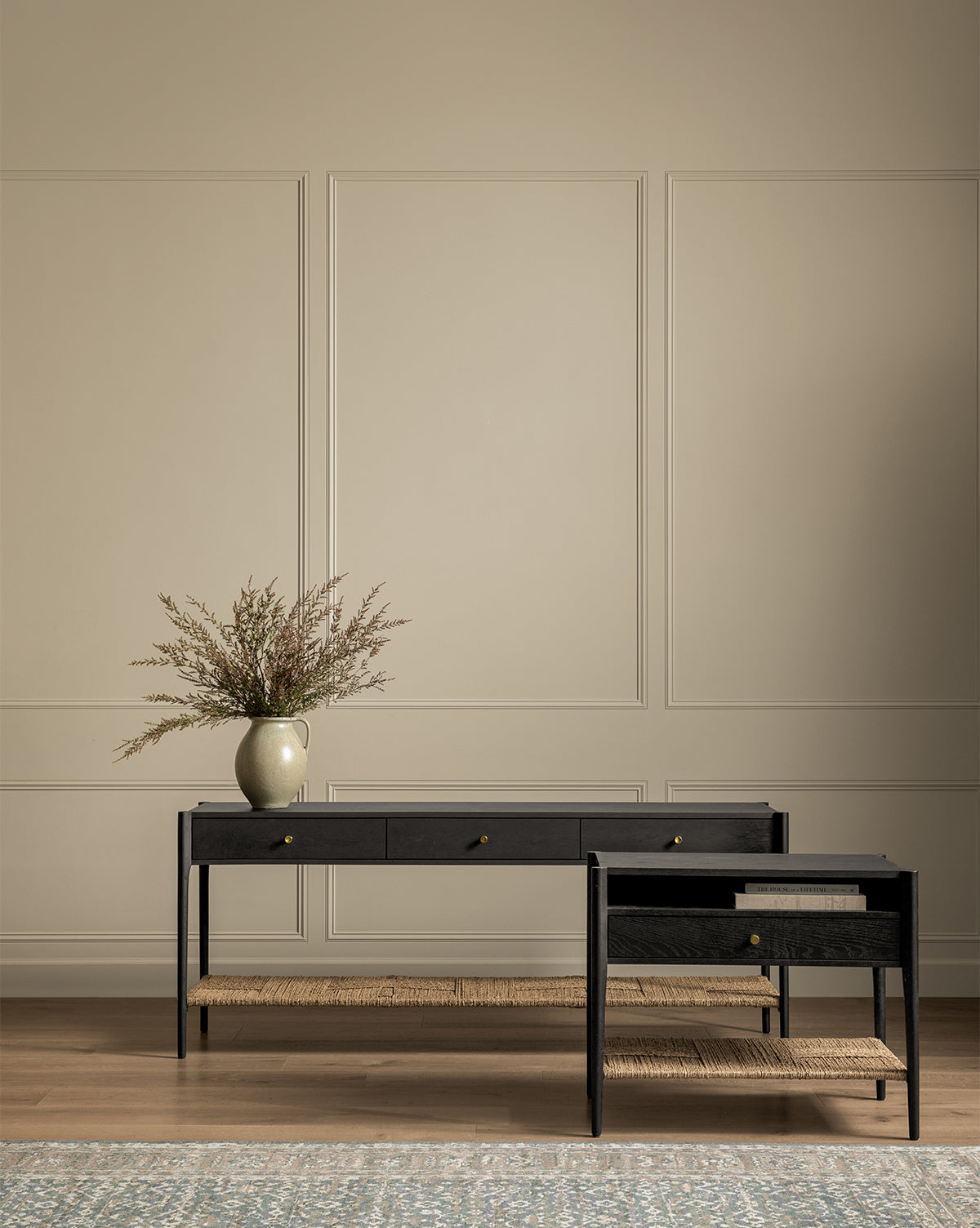 A minimalist room with beige paneled walls features the Dana Console in oak with brass hardware, topped with a ceramic vase and dried branches. A matching side table with woven shelves and a blue patterned rug complete the space by McGee & Co.