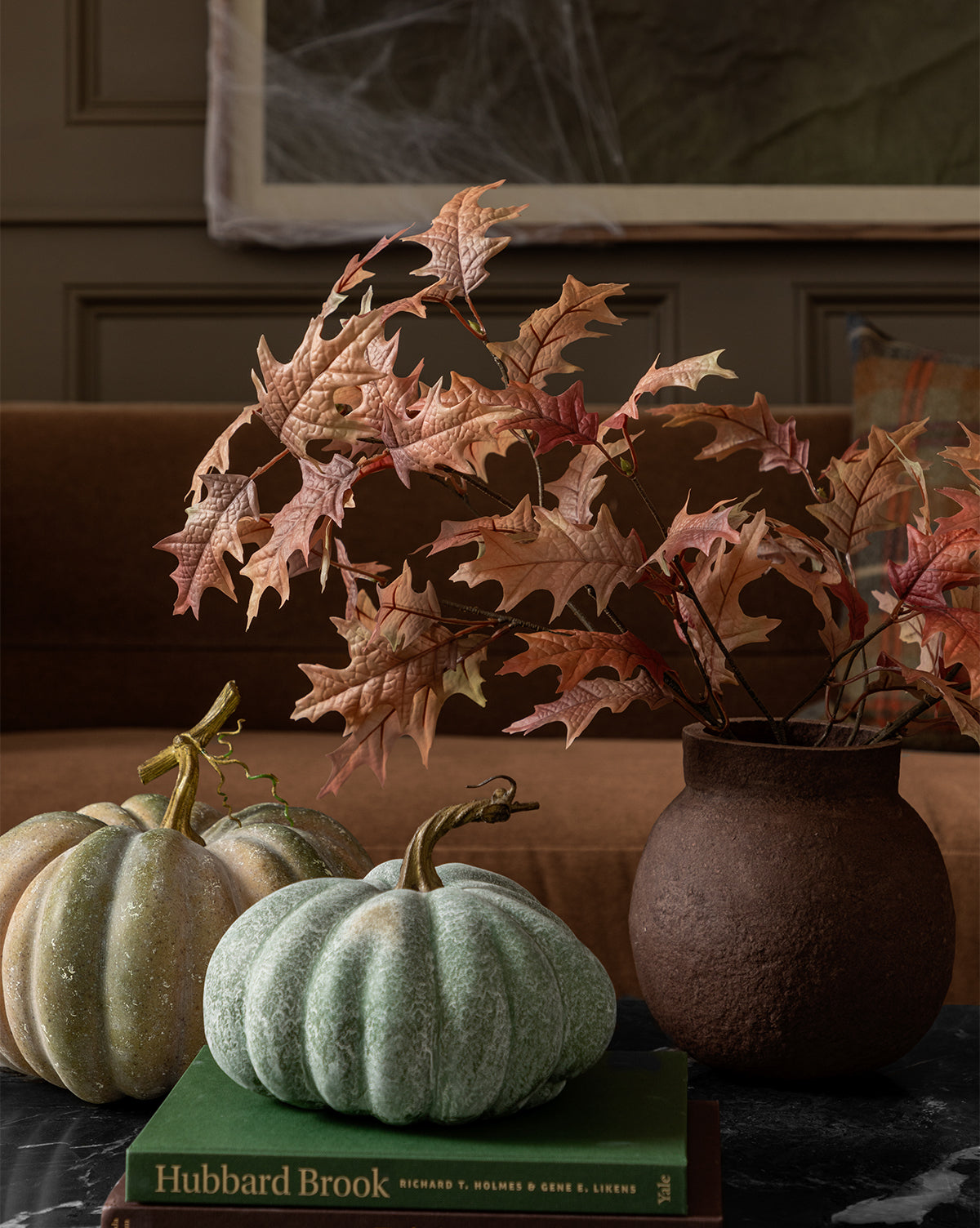 A Faux Autumn Oak Stem in a brown vase is on a table beside two green pumpkins and the book Hubbard Brook. A brown sofa and wall decorated with cobwebs create a cozy autumn backdrop.