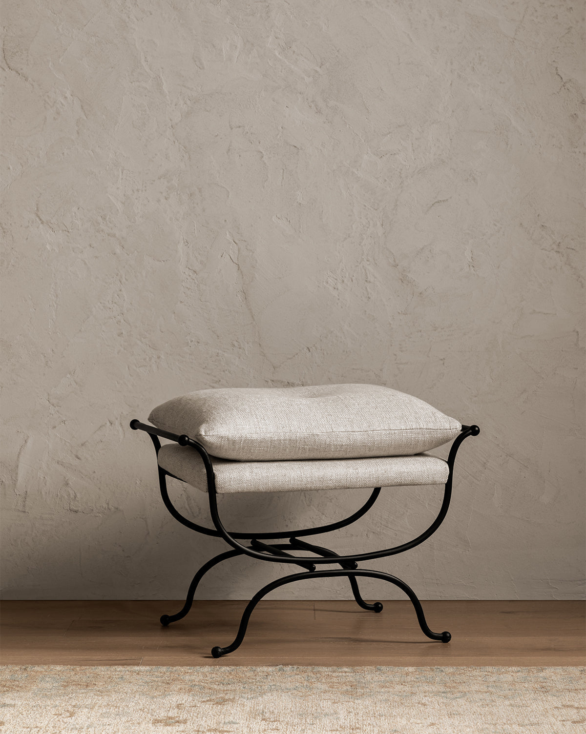 The Makerspalm Alameda Stool has beige linen upholstery and a curved matte black iron base. It stands on a wooden floor by a textured light gray wall, with part of a beige rug in the foreground. McGee & Co.