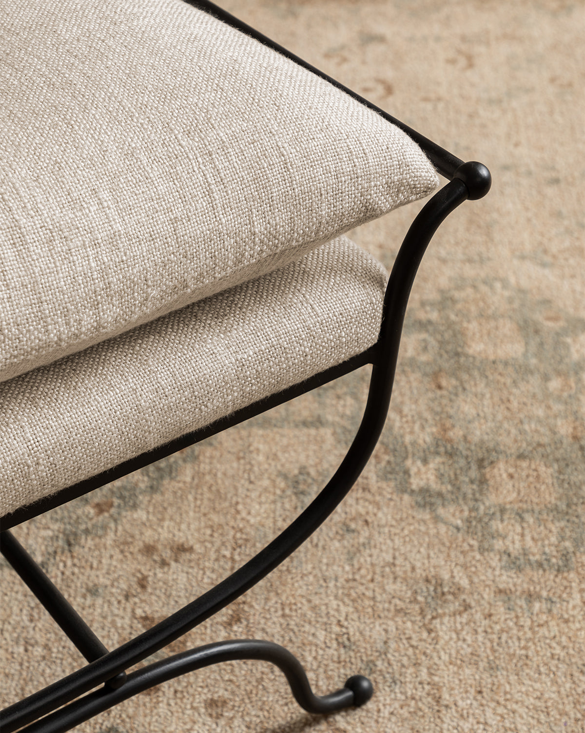 A close-up of the Makerspalm Alameda Stool, highlighting its matte black iron base and light beige cushion, placed on a neutral patterned rug by McGee & Co.