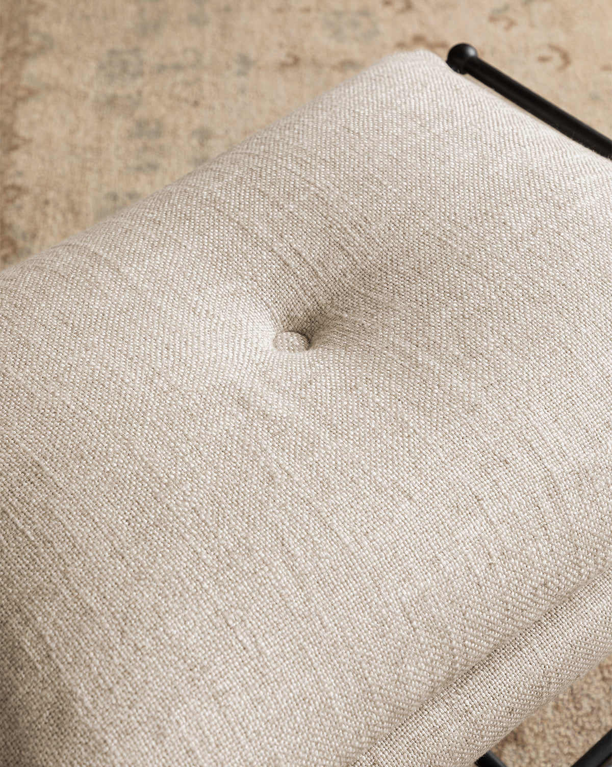 Close-up of a beige, textured linen cushion with a centered tuft on the Makerspalm Alameda Stool. The matte black iron base peeks out in the corner, resting on a patterned tan rug. McGee & Co.
