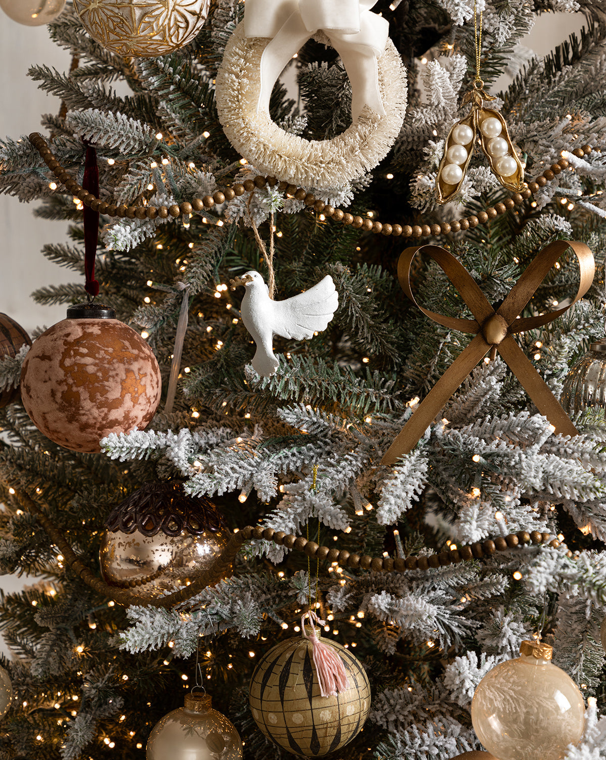 A Christmas tree with elegant neutral tones is adorned with Pearl Glass Baubles (Set of 21 Assorted), gold and white ornaments, a white dove, a gold ribbon bow, a wreath, and string lights among snow-frosted branches, by McGee & Co.