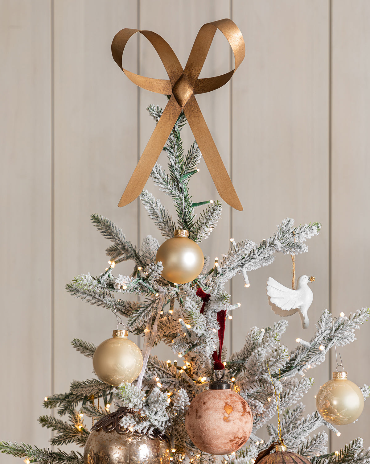 A Christmas tree adorned with gold and metallic ornaments, a set of 21 assorted Pearl Glass Baubles, a white dove ornament, and warm white string lights. Frosted branches and neutral tones contrast the light wood background, by McGee & Co.