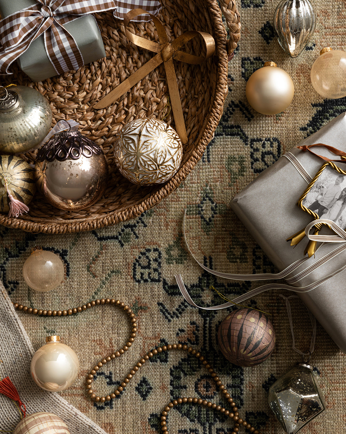 A woven basket of Christmas ribbon sits on a patterned rug, surrounded by wrapped gifts, gold beads, and the Pearl Glass Baubles (Set of 21 Assorted) in elegant gold, silver, and neutral holiday tones, by McGee & Co.