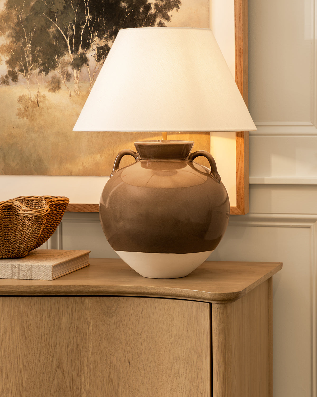 An Amphora Table Lamp in brown and beige ceramic with a white shade is displayed on a light wood cabinet beside a woven basket and book, with a landscape painting hanging on the wall behind, by McGee & Co.