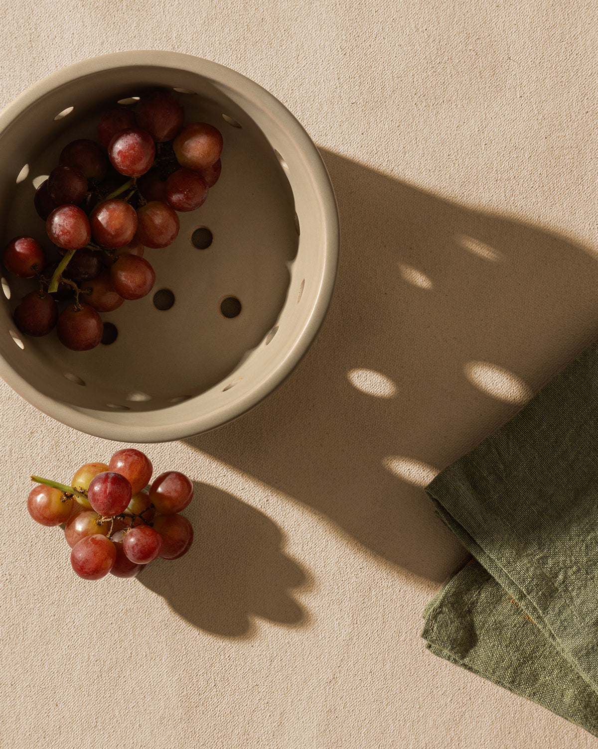 A beige Coen Berry Bowl with a matte finish holds red grapes on a light textured surface. A small bunch sits beside the stoneware bowl, near a folded green napkin. Both the Coen Berry Bowl and grapes cast distinct shadows by McGee & Co.