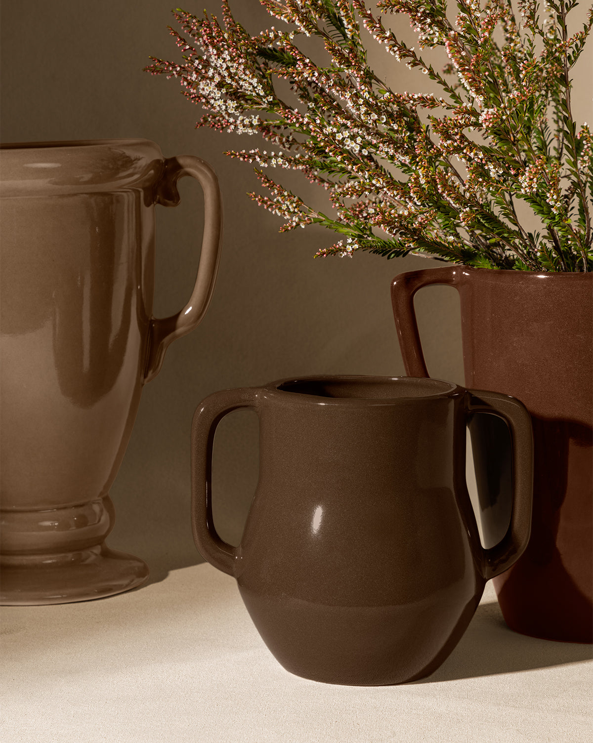 The Victoire Vase, an elegant earthenware vase in earthy brown tones, is displayed with two ceramic mugs on a neutral surface. One mug contains green foliage with white and pink flowers, creating a warm, cozy atmosphere under soft lighting, by McGee & Co.