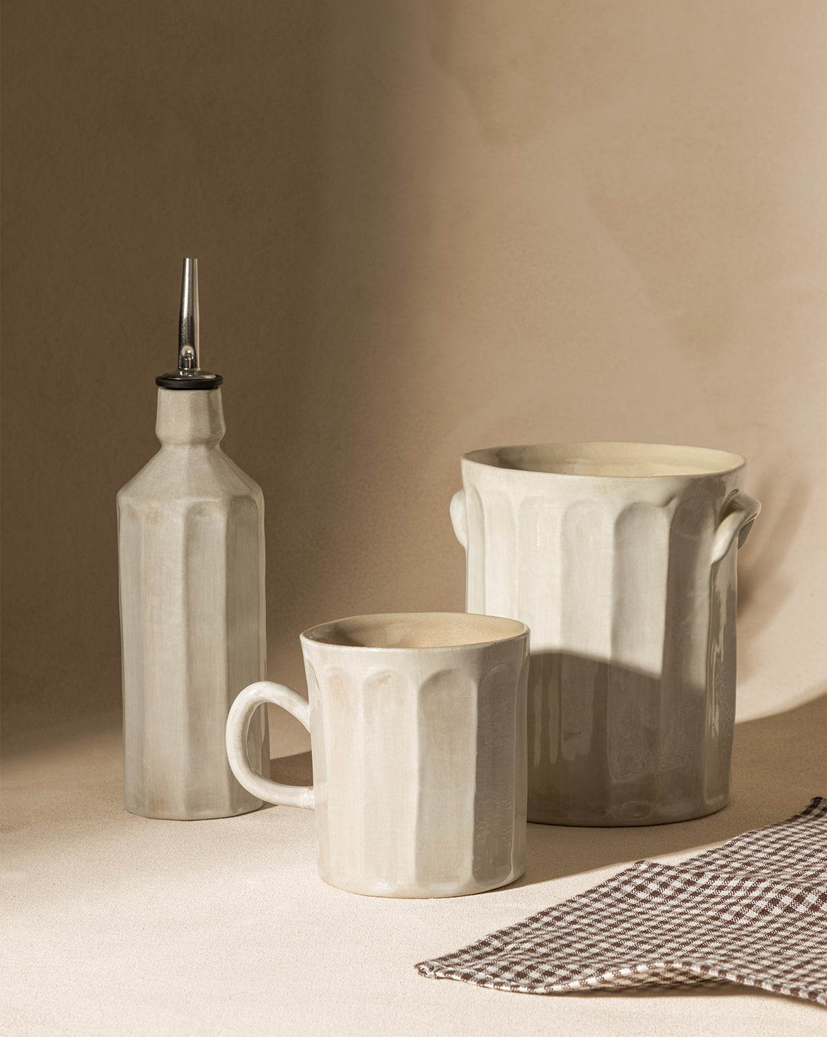 The Emilia Crock—a beige ceramic canister with a handle—sits on the kitchen counter beside a spouted bottle and handled mug. Nearby, a checkered cloth lies as soft sunlight creates gentle shadows across the scene.