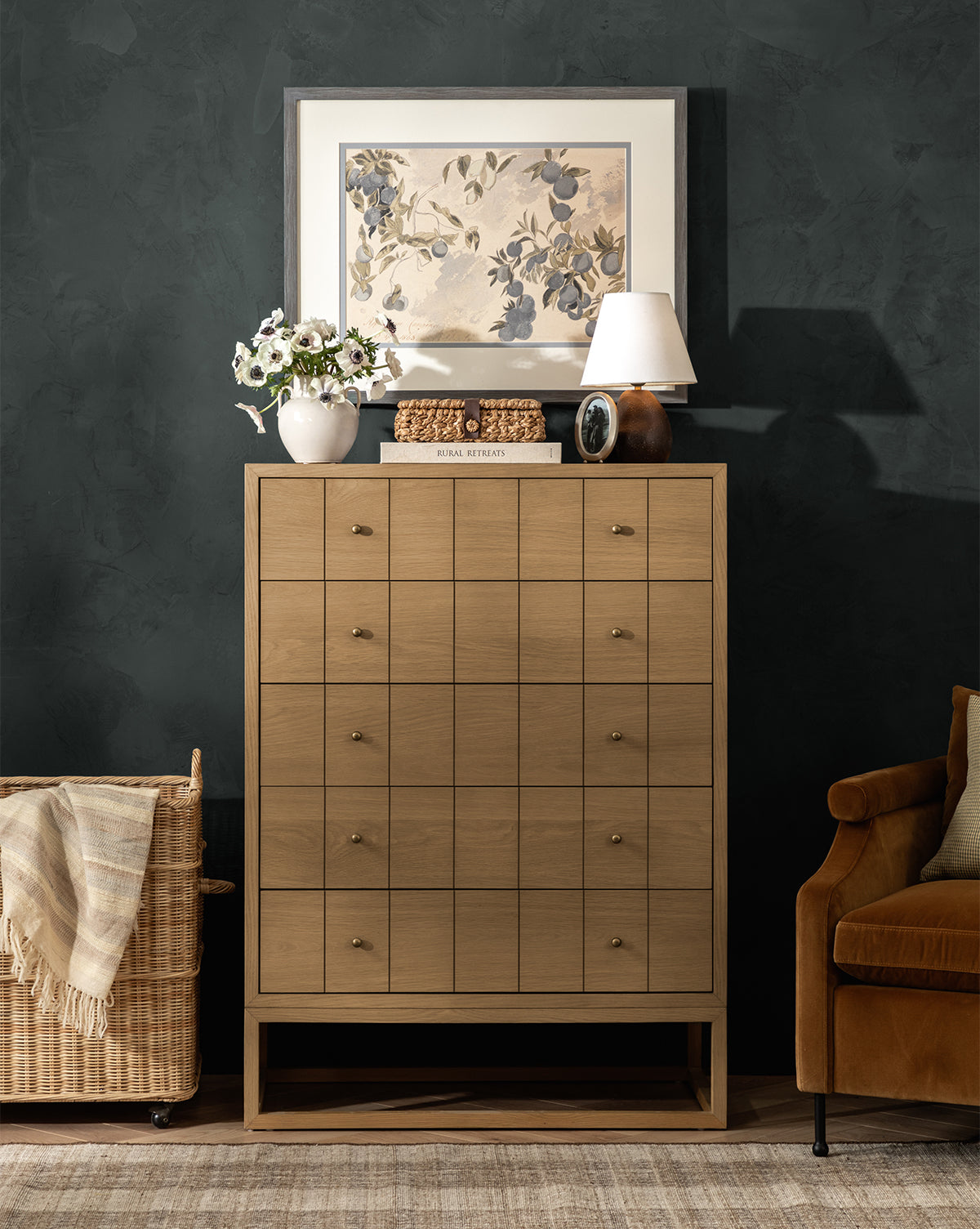 A light wood chest with twelve drawers stands against a dark wall. On top are a lamp, books, a white vase with flowers, decor, and Blue Fruits. Above hangs botanical art in a charcoal woodgrain frame. A wicker basket and brown armchair are nearby, by McGee & Co.