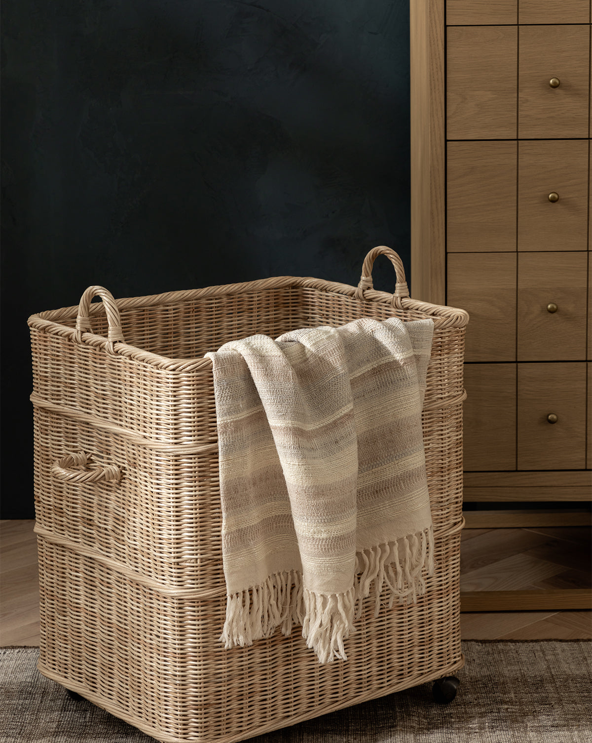 A woven wicker laundry basket with wheels and handles sits on a wood floor. The Laney Stripe Throw by TAL HANDWOVEN CLASSICS LLP, a beige, striped, fringed luxury blanket, hangs over the side. A wooden dresser with brass knobs is behind it. McGee & Co.