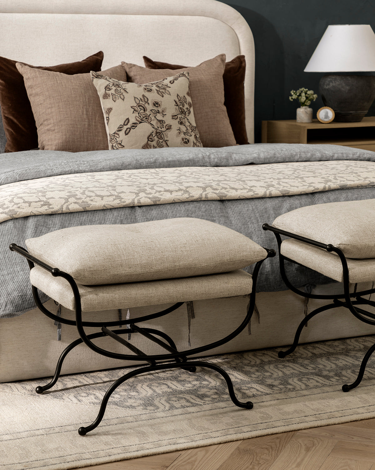 A cozy bedroom features a beige upholstered bed, layered brown and patterned pillows, a gray blanket, two Makerspalm Alameda Stools with matte black iron bases, plus a nightstand with a lamp and a decorative rug on the wooden floor, all curated in the inviting style of McGee & Co.