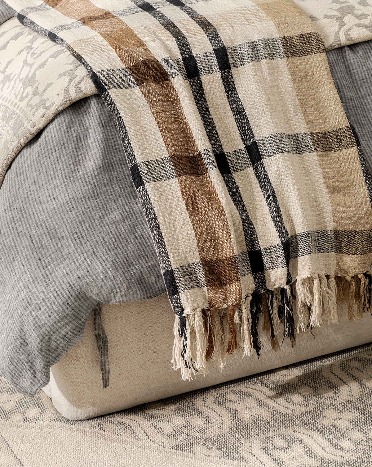 A close-up of a bed features layered bedding, including the Raj Albright Plaid Throw—a cozy cotton accent with brown and black stripes, fringed edges, and draped over the foot for added texture. McGee & Co.