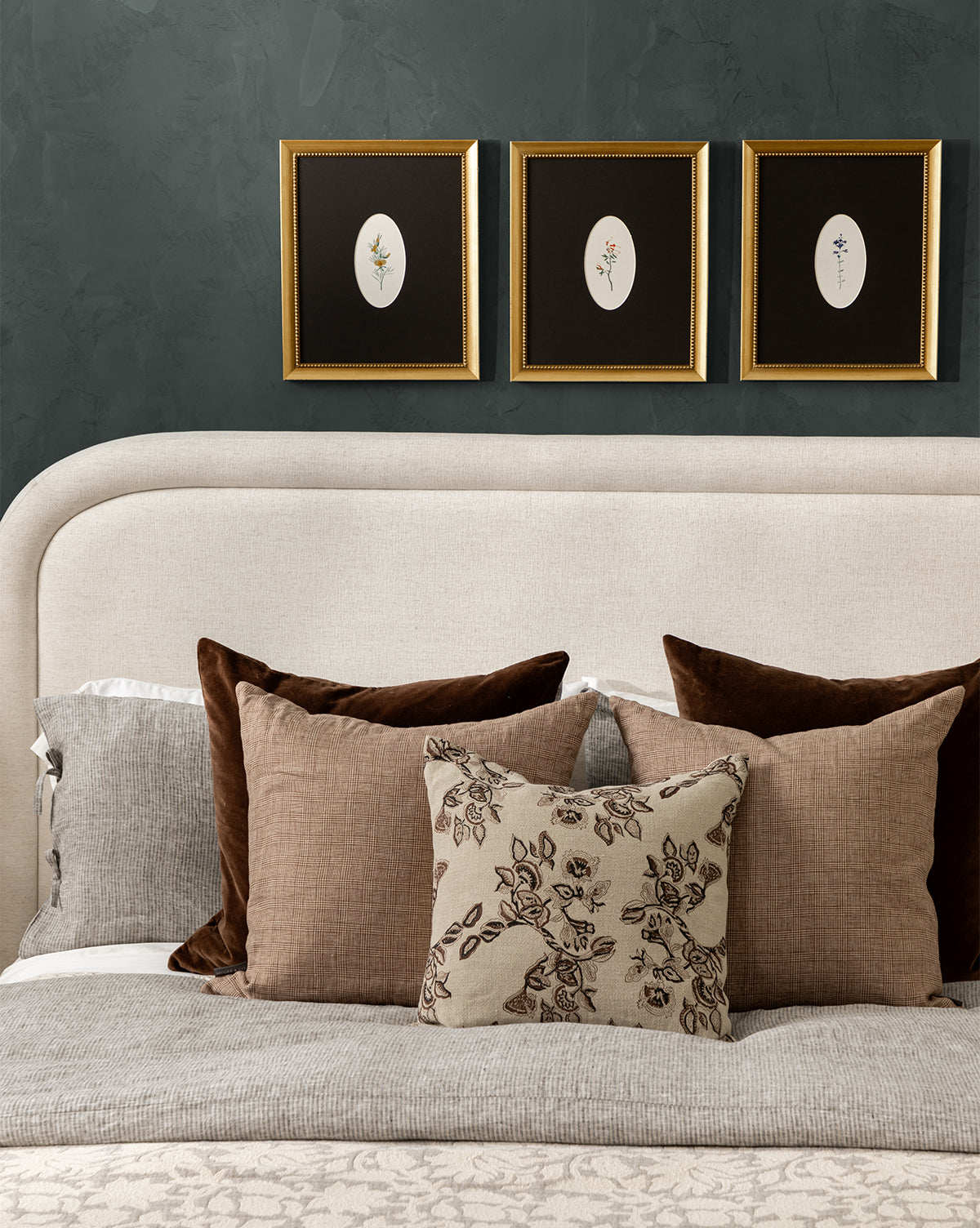 A neatly made bed with brown and beige pillows and a beige headboard sits against a dark green wall, topped with three Single Blooms 2 botanical prints in gold-finished wood frames arranged above the bed, by McGee & Co.