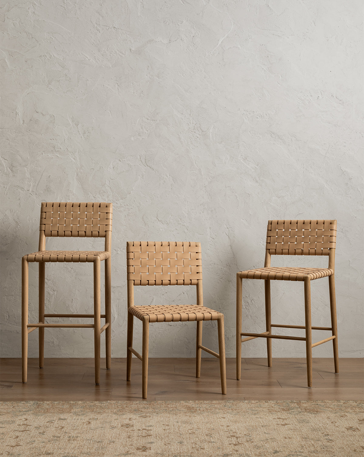 Three Greely Stools with white oak frames and woven seats, in varying heights, are arranged side by side against a textured light gray wall on a wooden floor, with part of a beige rug in the foreground by McGee & Co.