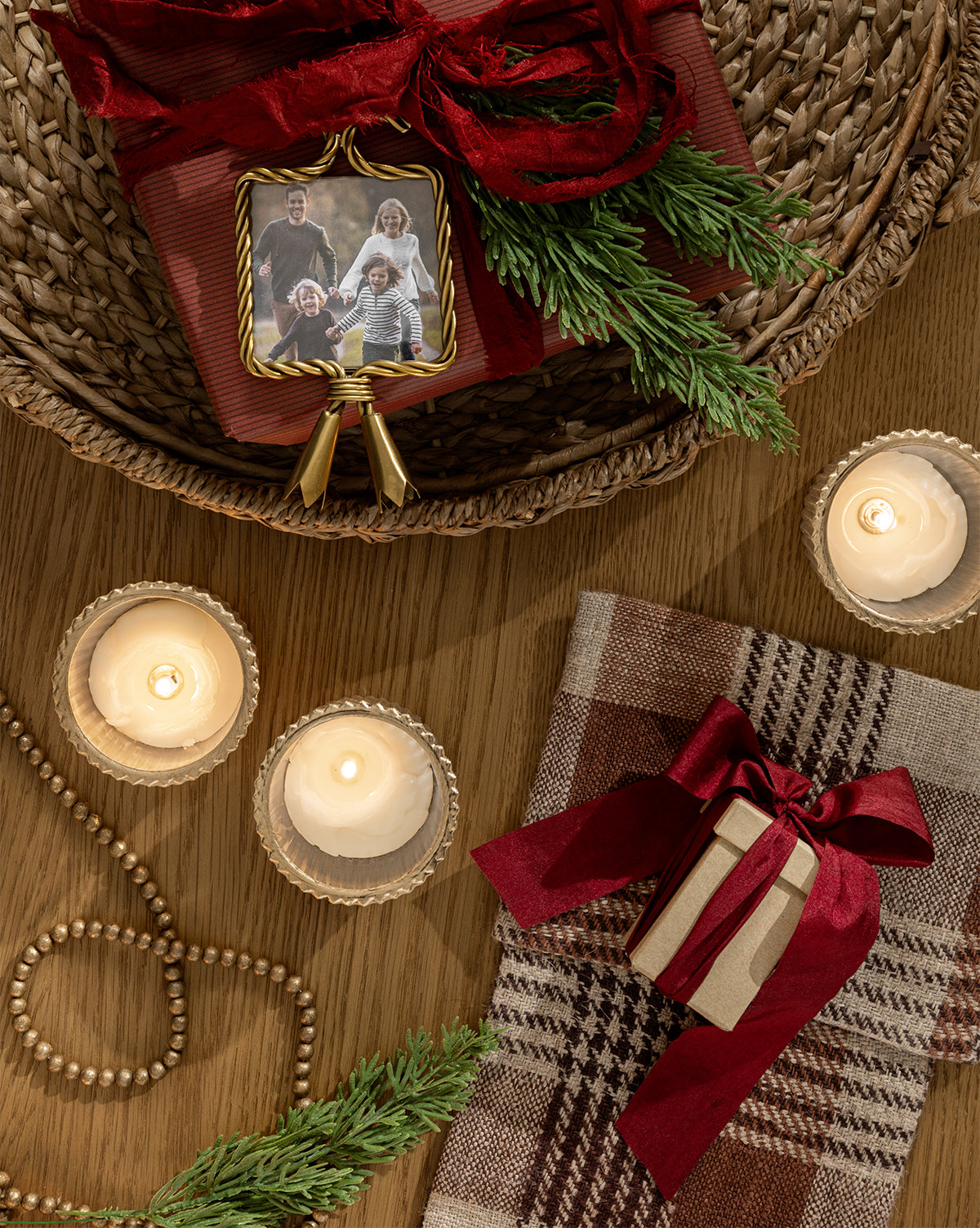 A festive scene with candles, a wrapped gift box, evergreen sprigs, plaid fabric, wooden beads, a framed family photo, and the Tassel Picture Frame Ornament displayed on a wooden table, by McGee & Co.