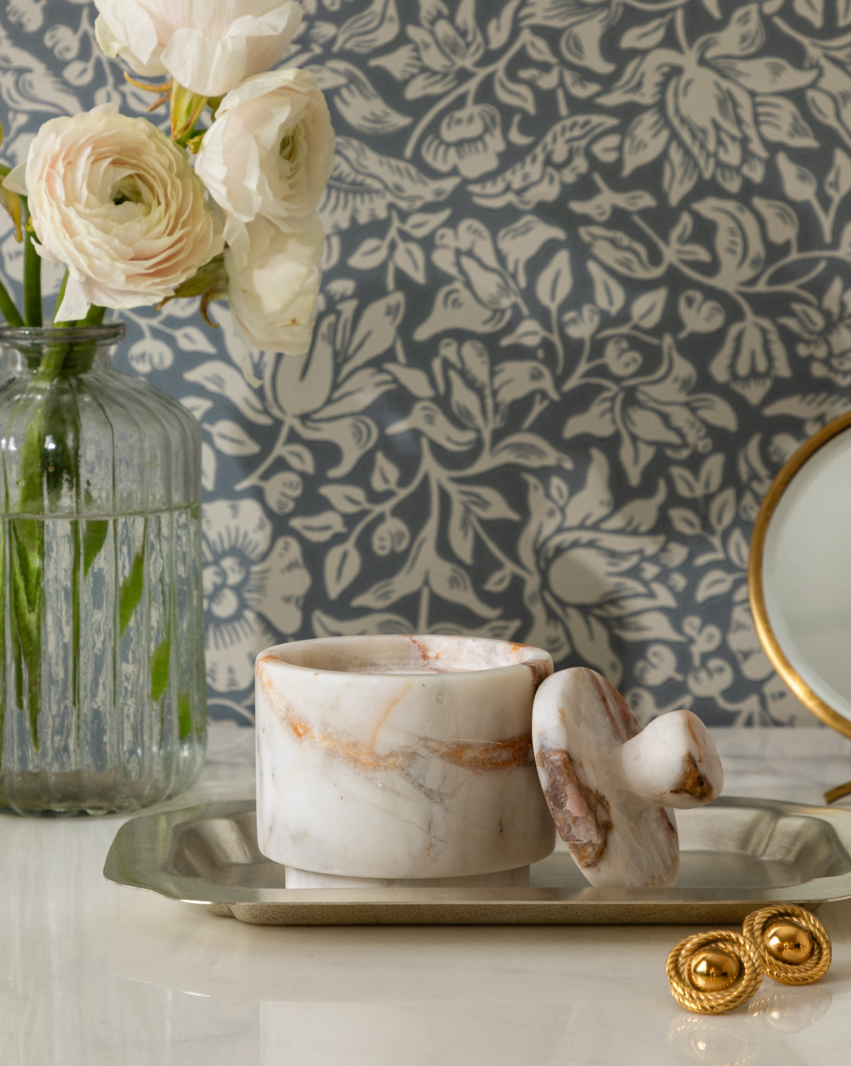 A marble mortar and pestle sit on a tray beside gold earrings and the Verona Matte Marble Lidded Jar, with a glass vase of white flowers and floral-patterned wallpaper in the background, by McGee & Co.