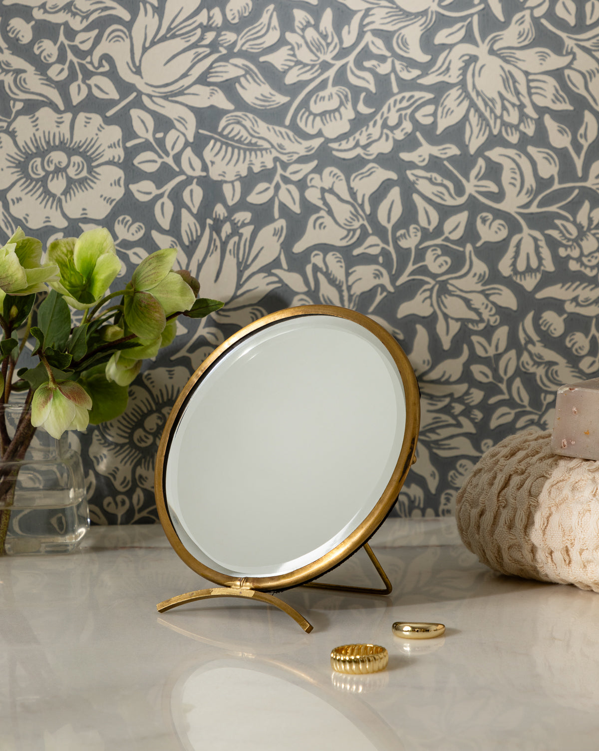 The Antique Brass Folding Mirror stands on a marble countertop beside a potted plant, a textured towel, gold jewelry, and pink soap, all set against floral-patterned wallpaper, by McGee & Co.