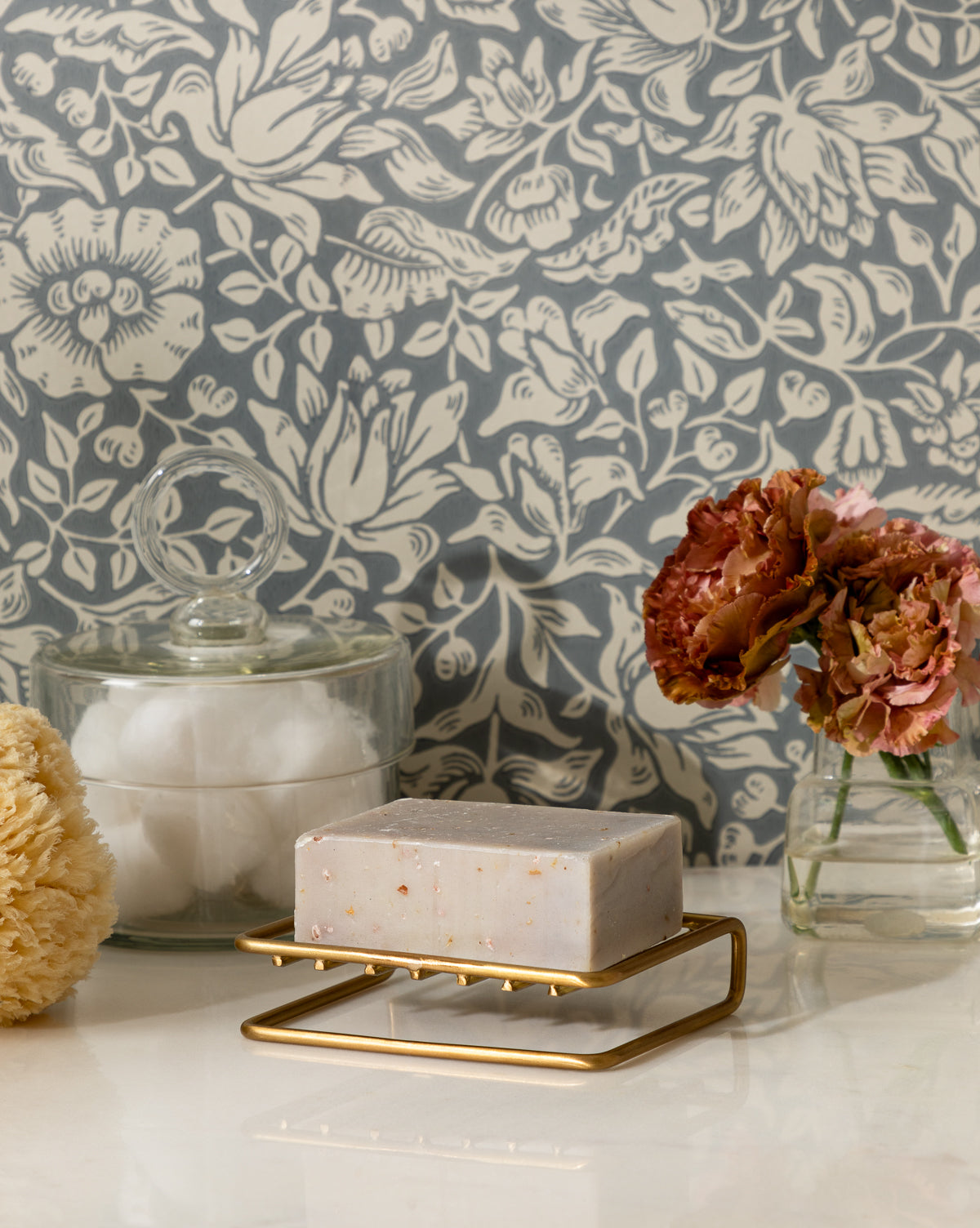 A Brass Soap Dish holds a bar of soap beside a glass jar of cotton balls, a yellow sponge, and a small vase with pink flowers arranged on bathroom counters against floral-patterned wallpaper.