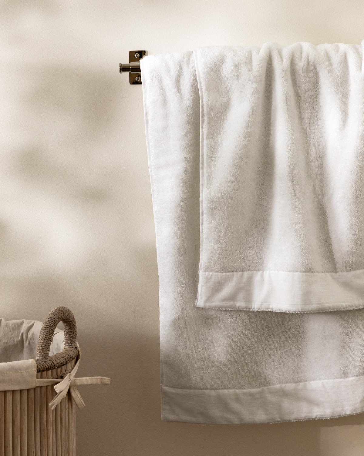 A towel from the Dedra Bath Collection hangs on a wall bar beside a beige woven laundry basket with a rope handle and bow, against a light-colored wall.