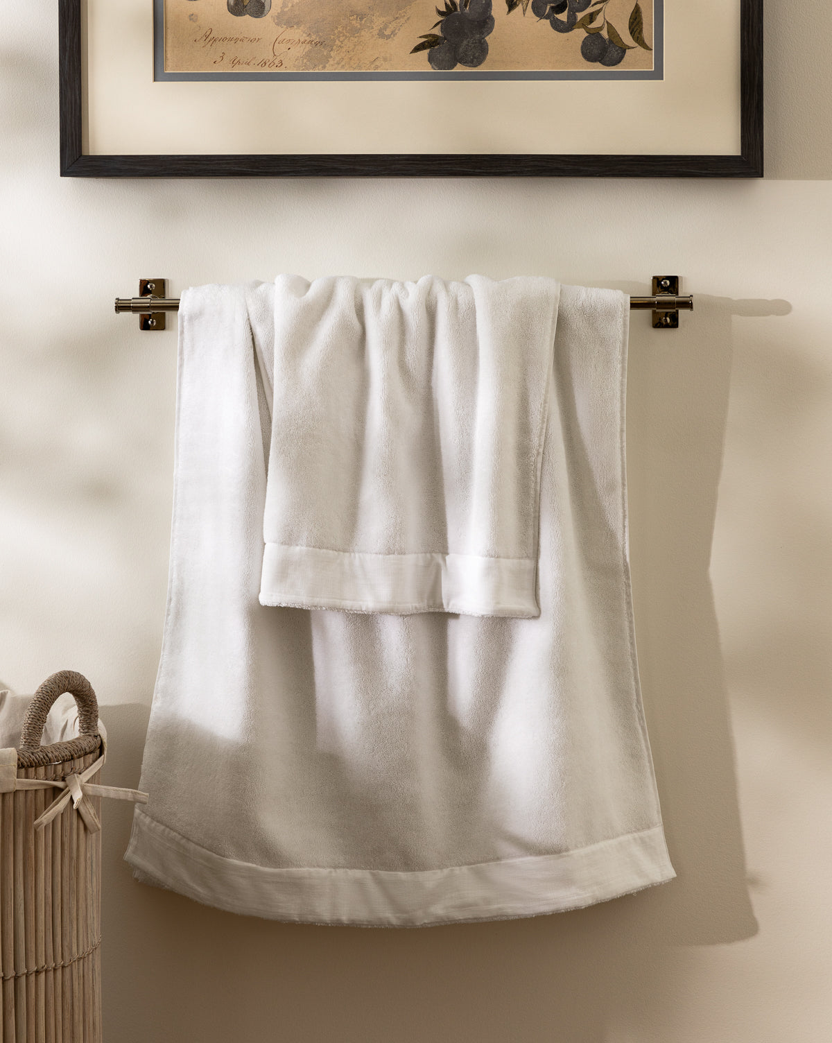 Towels from the Dedra Bath Collection hang on a silver rack against a beige wall, with framed art above and a wicker laundry basket partly visible to the left.