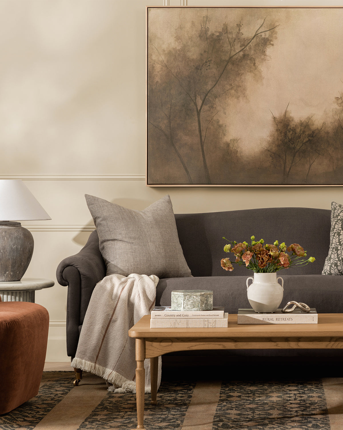 A cozy living room features a dark sofa with gray pillows, a light throw blanket, and a wooden coffee table topped with flowers, books, and a decorative box. Above the sofa hangs Restored by Brittany Schulz on museum-quality canvas, by McGee & Co.