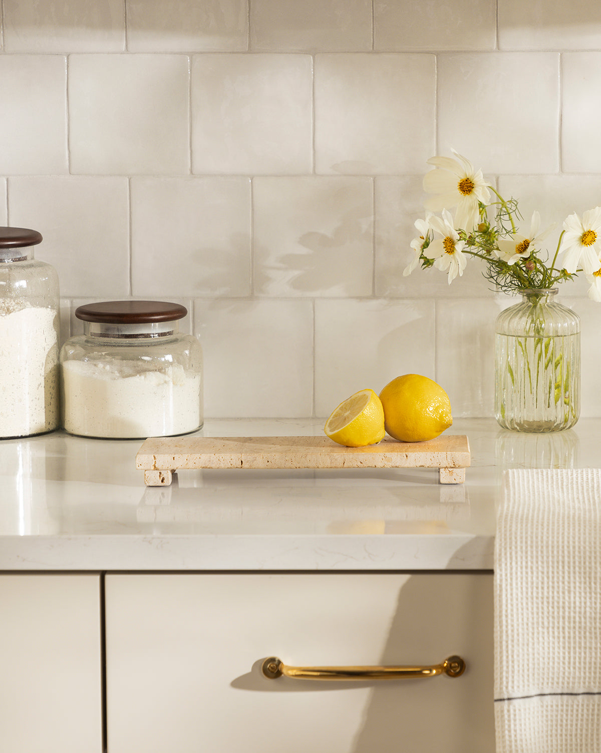 A kitchen counter displays a Beige Travertine Footed Serving Board with two lemons (one halved). Nearby are glass jars of flour, a jar of white flowers, and a hanging towel, all set against a tiled backsplash, by McGee & Co.