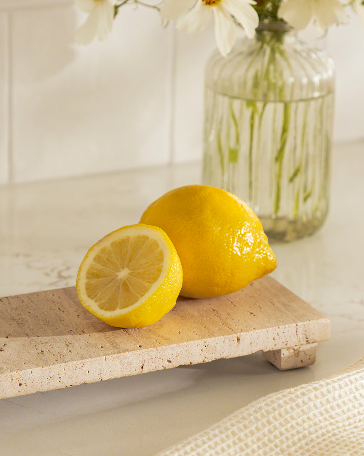 A whole lemon and a halved lemon sit on a Beige Travertine Footed Serving Board, with a glass vase of white flowers in the background on a light-colored countertop, by McGee & Co.