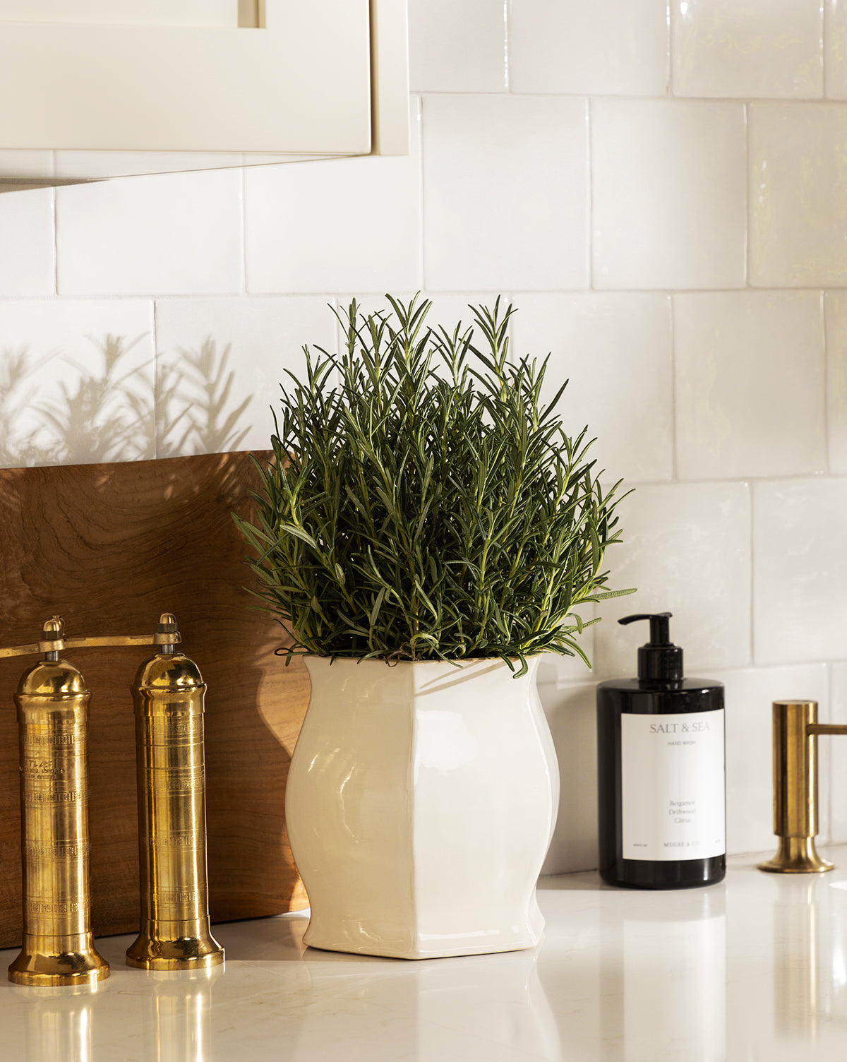 A rosemary plant in a Kasha Cream Pot sits on a kitchen counter beside gold salt and pepper mills, a soap dispenser, and a paper towel holder, with white tiled backsplash and a wooden cutting board in the background, by McGee & Co.
