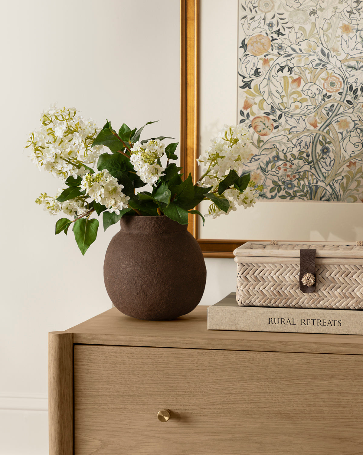 A Faux White Lilac Stem in a brown vase sits on a wooden dresser beside a woven box and the book Rural Retreats, with a framed botanical print hanging on the wall behind them.