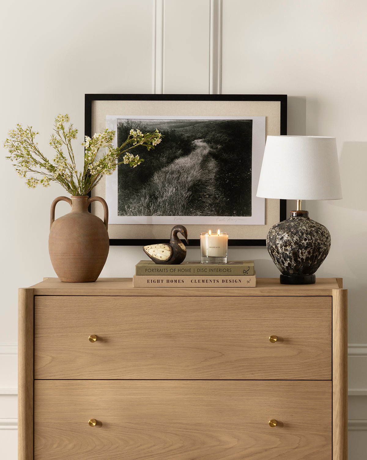 A light wood dresser holds a vintage-inspired ceramic vase with flowers, two stacked books, a small bird figurine, a lit candle, the Zella Swan Object, a marble-patterned lamp, and a framed landscape photo leaning against the wall.