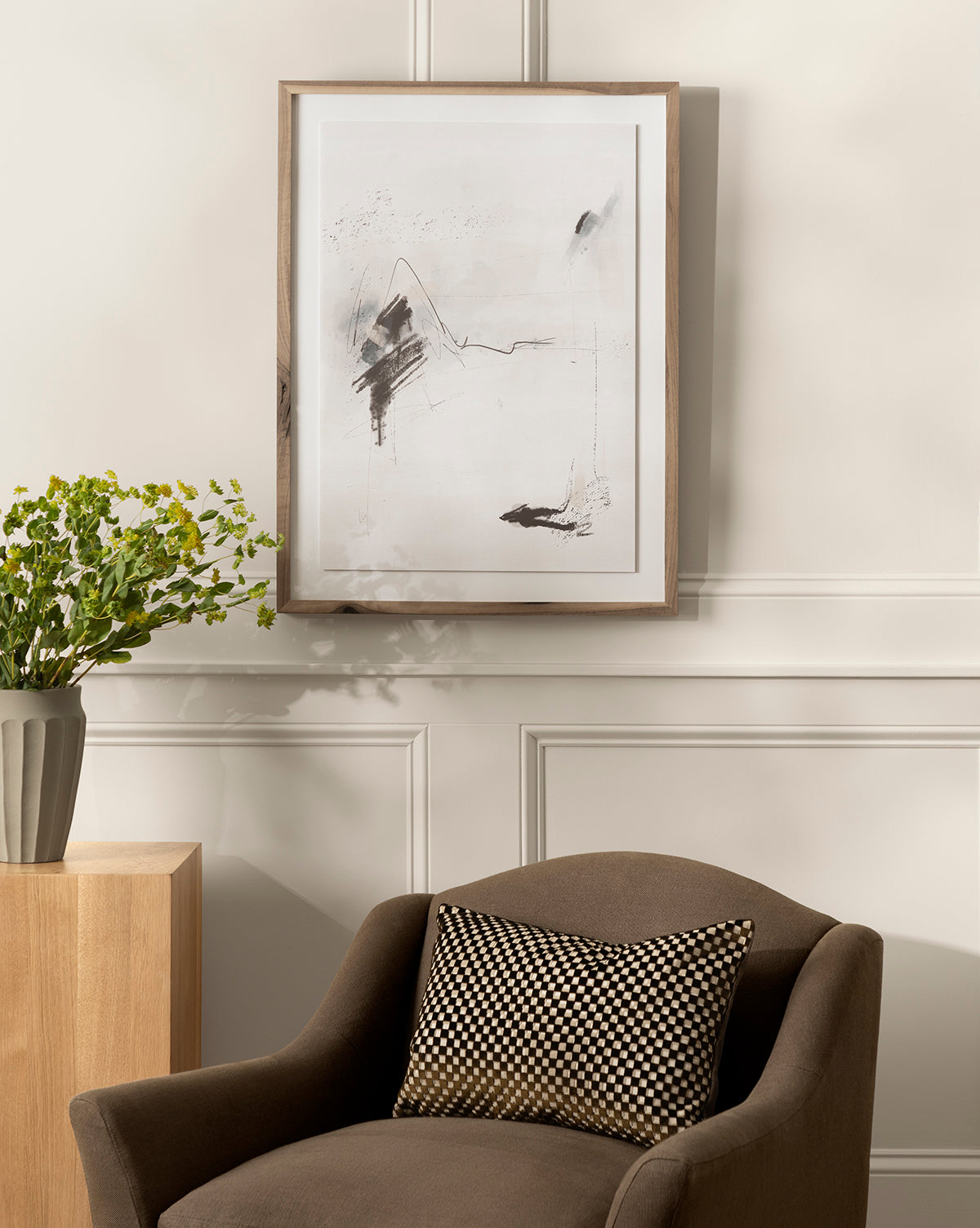 A modern living space features a brown armchair with a patterned cushion, a wooden side table with a vase of green foliage, and Momento II by Coup dEsprit in a rustic walnut frame on a light-paneled wall.