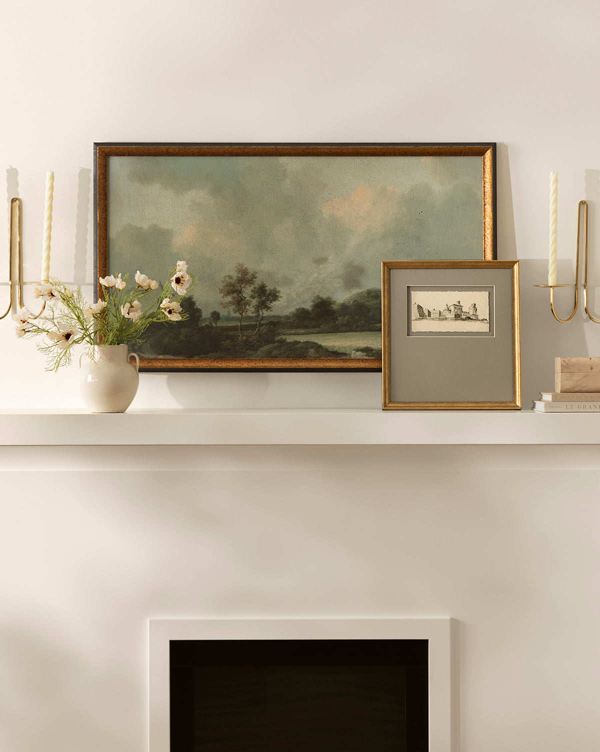 A mantel with vintage wall art, including Clouds Above a Field, a smaller framed sketch, brass candle sconces with twisted candles, a vase of flowers, and a stack of books set against a light-colored wall.