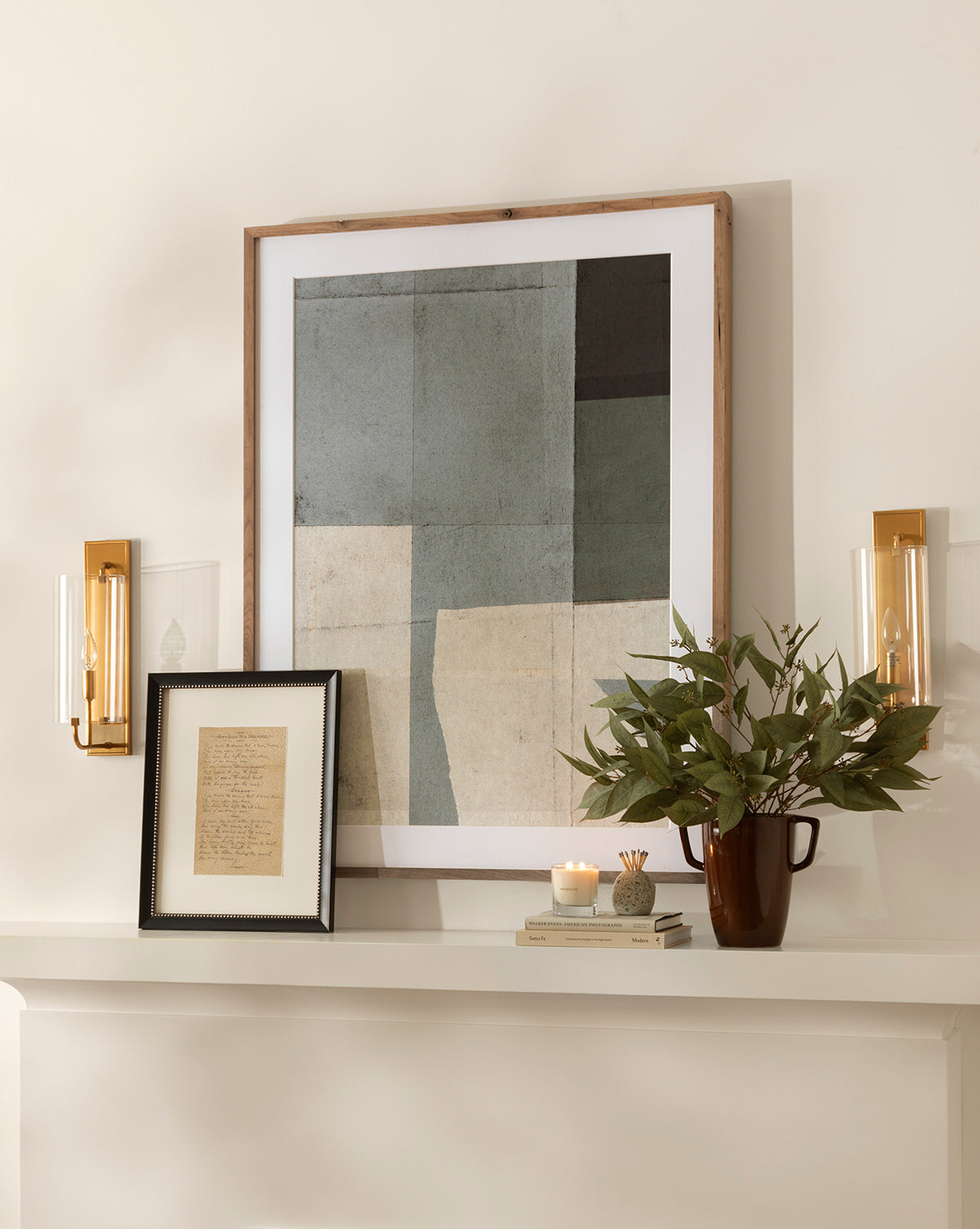 A minimalist mantel displays a modern abstract painting, a framed handwritten note, two brass sconces, the Faux Eucalyptus Seed Stem in a brown vase, small candles, and stacked books—all set against a light-colored wall.