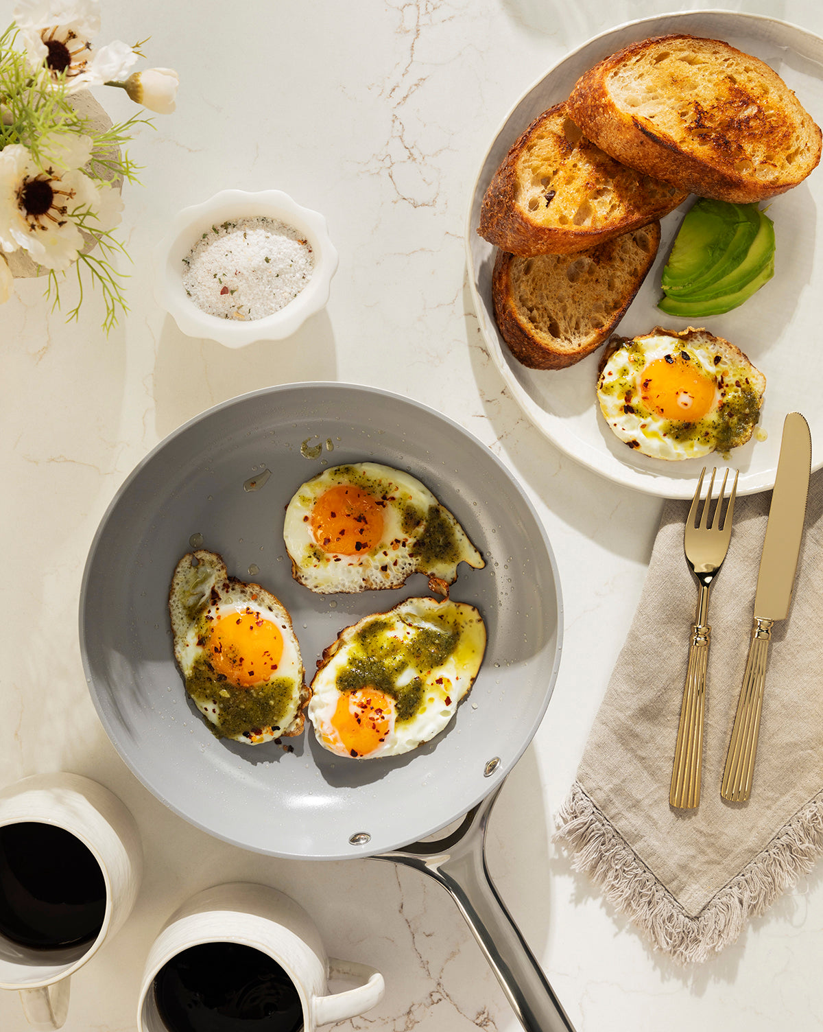 A frying pan with three sunny-side-up eggs topped with green sauce rests on a white countertop, next to toasted bread, avocado, coffee, utensils, a white napkin, and a Scalloped Marble Bowl filled with salt by McGee & Co.