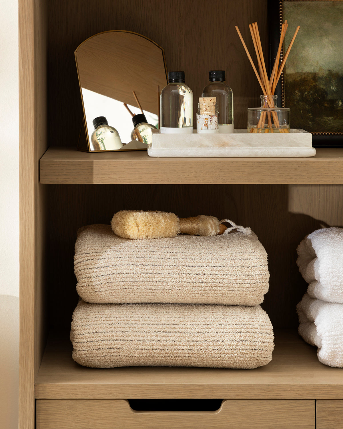 A wooden shelf showcases beige towels, a natural bristle brush, mini toiletries, a glass jar, reed diffuser sticks, and the Standing Vanity Mirror with a brass finish, creating an organized and spa-like bathroom countertop display.