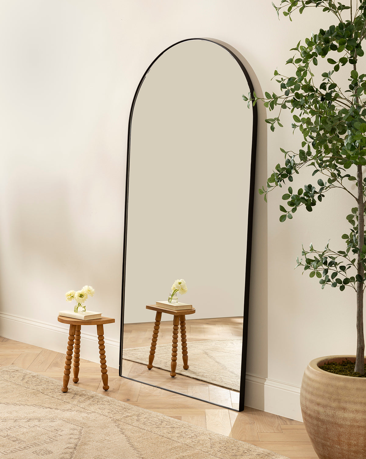 The Gala Floor Mirror, featuring a large arched silhouette and black frame, leans against a light wall. A small turned-leg table with a vase of white flowers and a nearby potted plant complete the look on the wooden floor by McGee & Co.