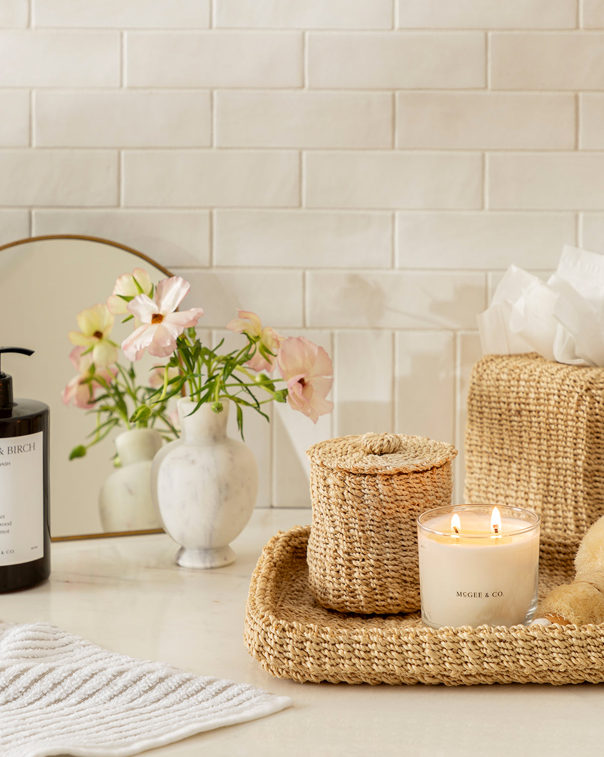 A cozy bathroom counter displays the Deryn Woven Tray with a lit candle, plus woven baskets, marble vase of pink flowers, striped towel, tissue box, and soap dispenser—all arranged before a tile backsplash and small mirror.
