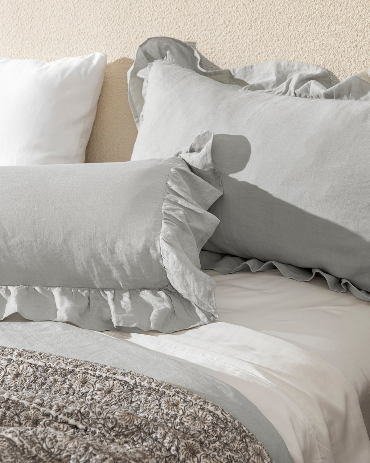 A neatly made bed with light gray ruffled pillows, Ruffle Linen Shams (Set of 2), white sheets, and a textured gray blanket creates romantic bedding against a beige wall.