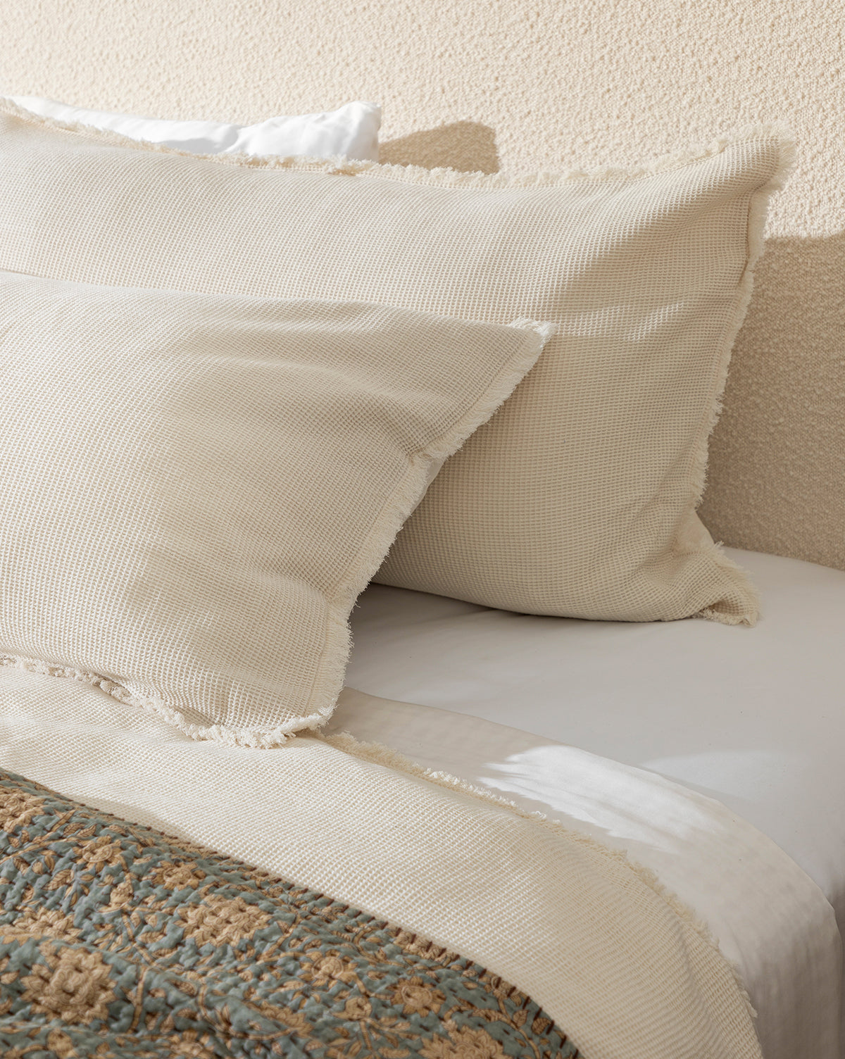 Two cream-colored Helene Waffle Shams rest against a beige wall on a neatly made bed, complemented by a cream throw, patterned blanket, and neutral white bedsheet.