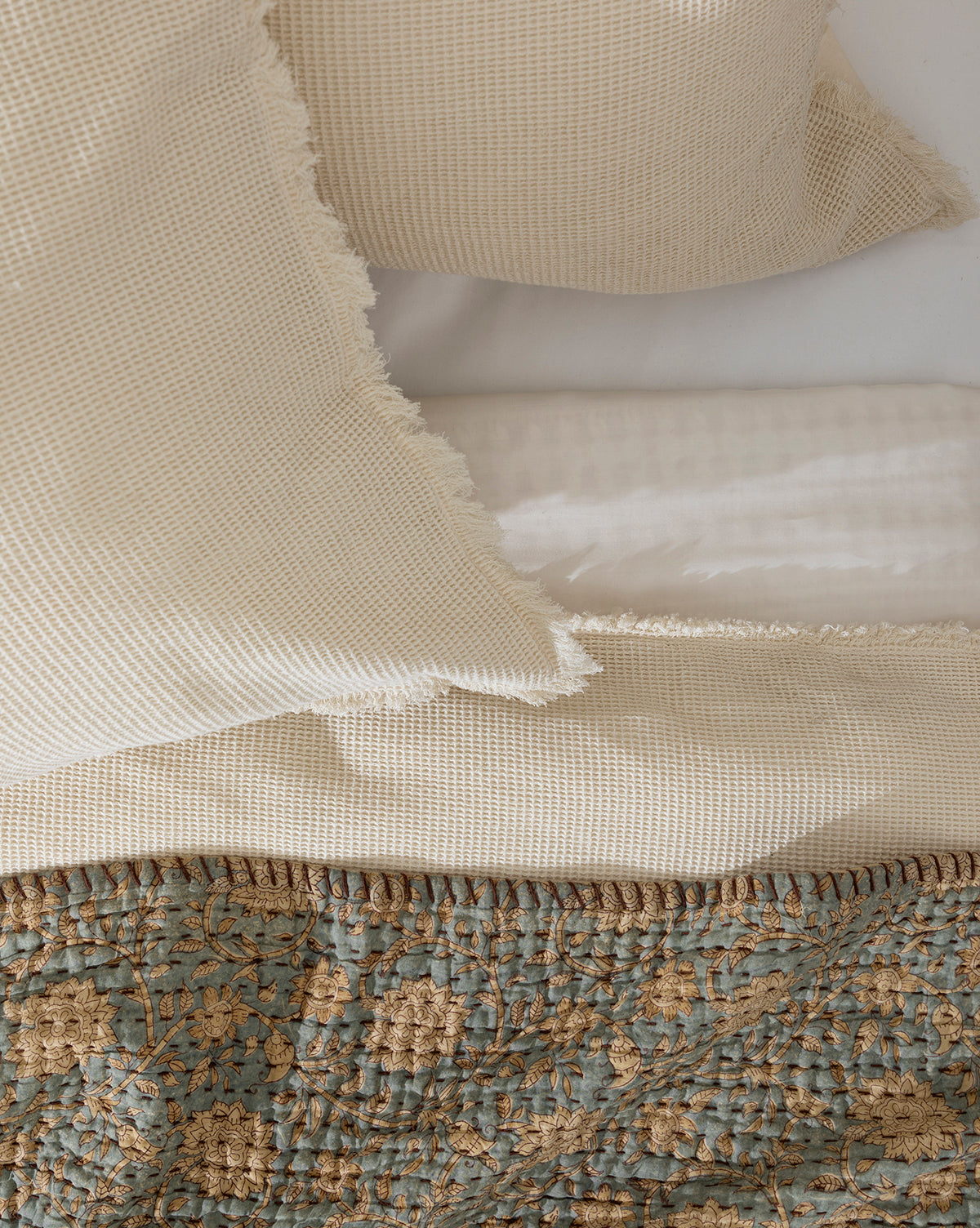 Cream-colored textured pillows and a matching blanket rest on a white sheet, with the Gabriella Block Print Quilt in blue-green and gold floral pattern partially visible at the bottom of the image.