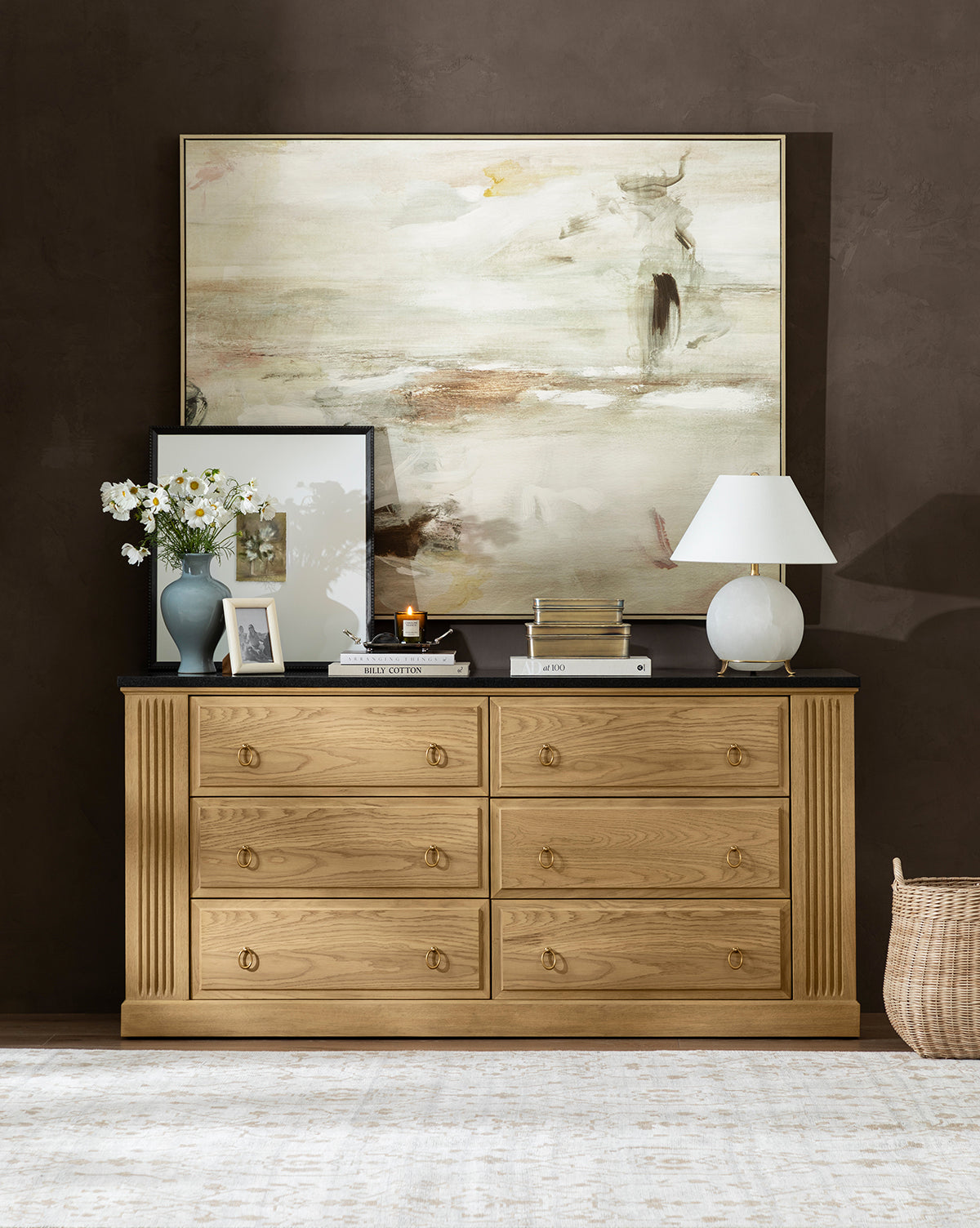 A wooden dresser with six drawers is adorned with books, a white lamp, flowers, candles, and stacked art frames. On the dark wall behind hangs the large gallery-wrapped canvas Neutral Abyss. A woven basket sits on the floor nearby, by McGee & Co.