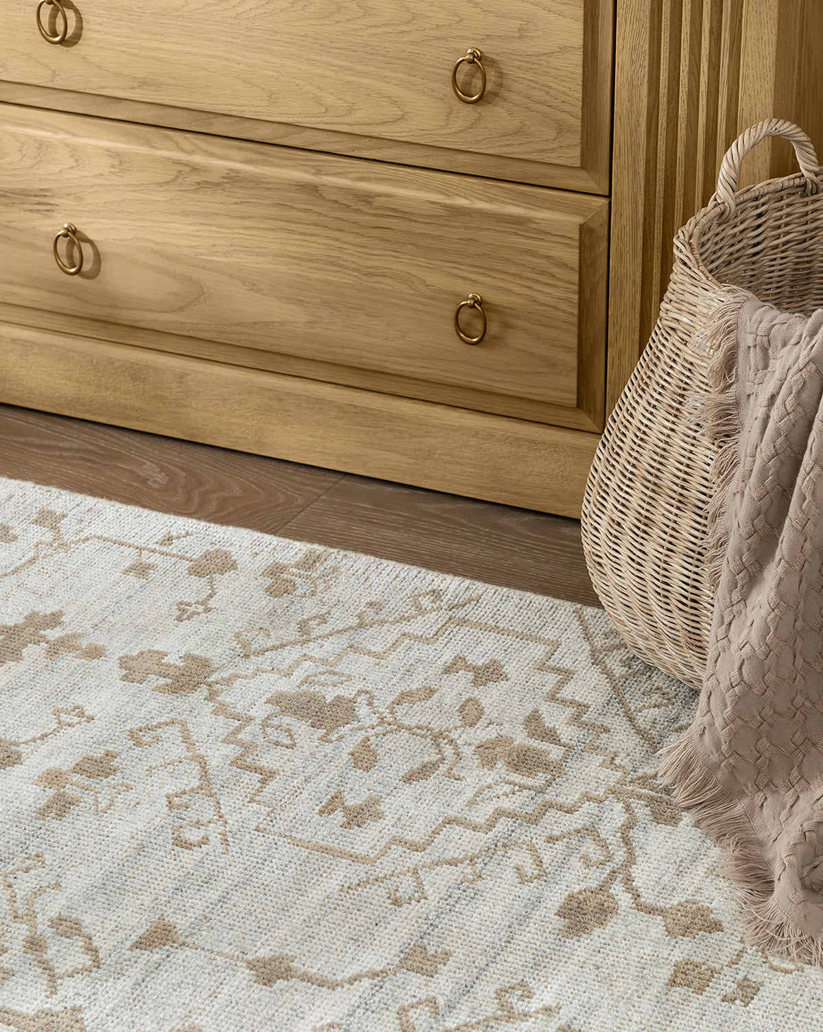 A close-up of the Lexington Hand-Knotted Wool Rug in a light pattern on a wood floor, next to a woven basket with a beige blanket, and in front of a light wood dresser featuring brass handles, by McGee & Co.