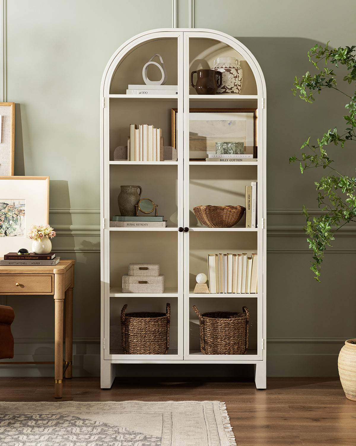The Brookes Cabinet, a white glass door cabinet, showcases books, baskets, vases, and decor. Placed by a green wall near a wooden desk with plants and framed art, it adds charm and coziness to the room by McGee & Co.