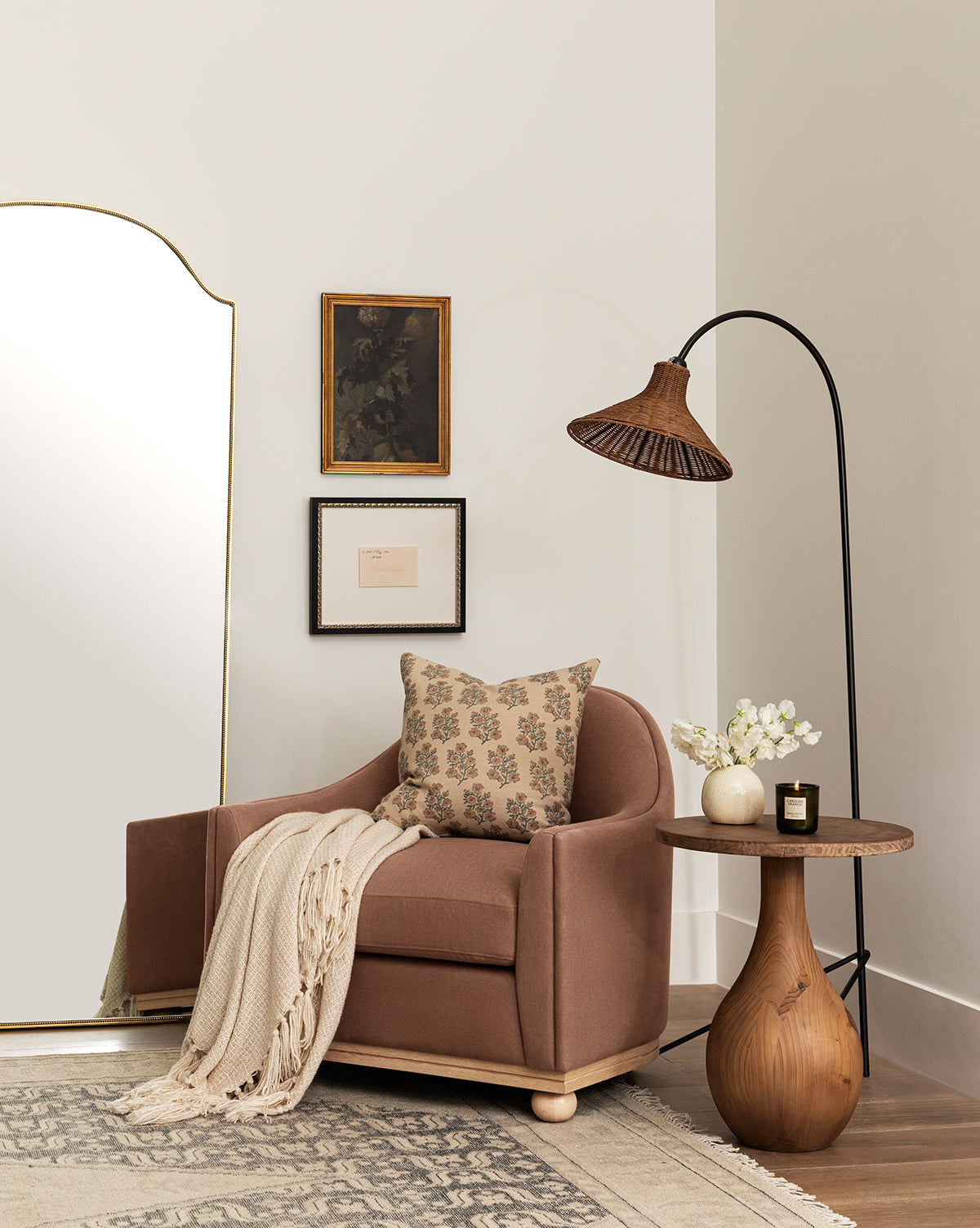 A cozy corner features a brown armchair, floral pillow, cream throw, and round wooden side table with flowers and a candle. The space is decorated with the Johanson Woven Floor Lamp, framed art, and a large mirror by McGee & Co.