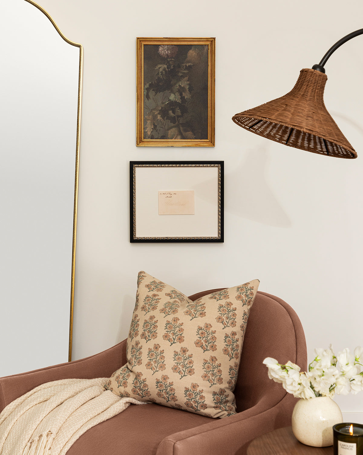 A cozy corner features a mauve armchair, floral pillow, and light throw. A wicker lamp hangs above vintage art in wood frames. A large mirror and vase of white flowers complete the decor, while Hard Cider adds a refreshing touch to the scene, by McGee & Co.