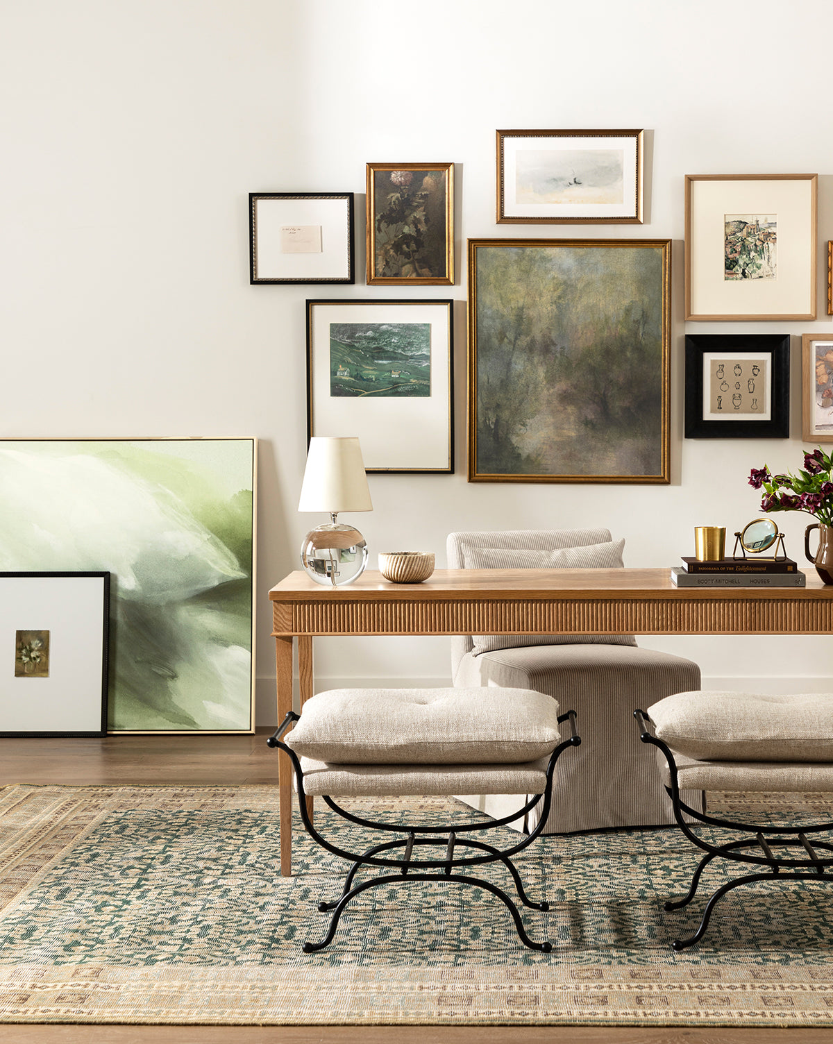 A stylish home office with a wood desk, neutral chair, two stools, a green-patterned rug, and the Fields of Green canvas art featured above the desk. Abstract art completes the gallery wall, while a table lamp and decor add finishing touches, by McGee & Co.