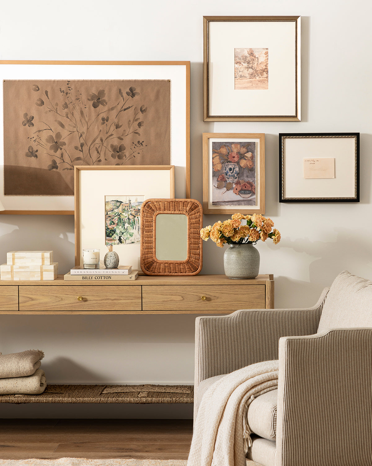 A cozy living room corner featuring a wood console table topped with Hard Cider, vintage art in distressed frames on a cream wall, a small mirror, books, yellow flowers, and a striped armchair with a blanket on the lower shelf by McGee & Co.