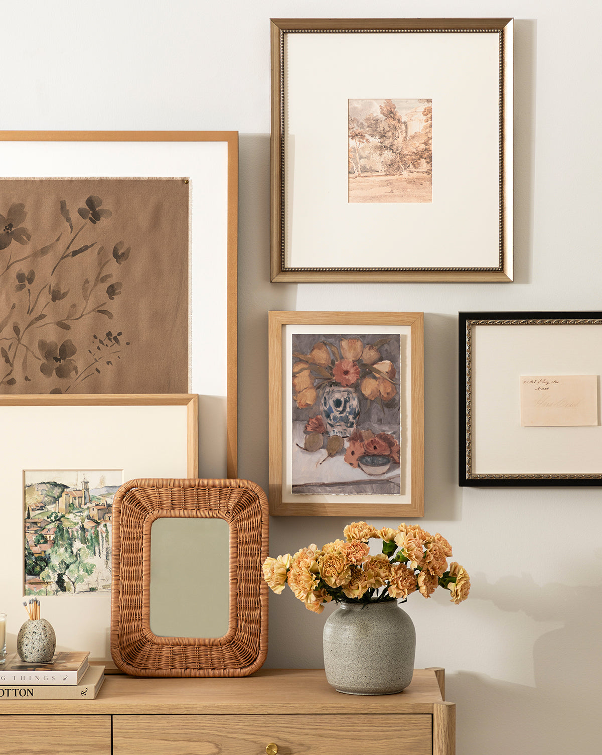 A wooden dresser with a vase of dried flowers and the small Jera Shelf Mirror sits below a gallery wall displaying framed landscape and floral artworks, by McGee & Co.