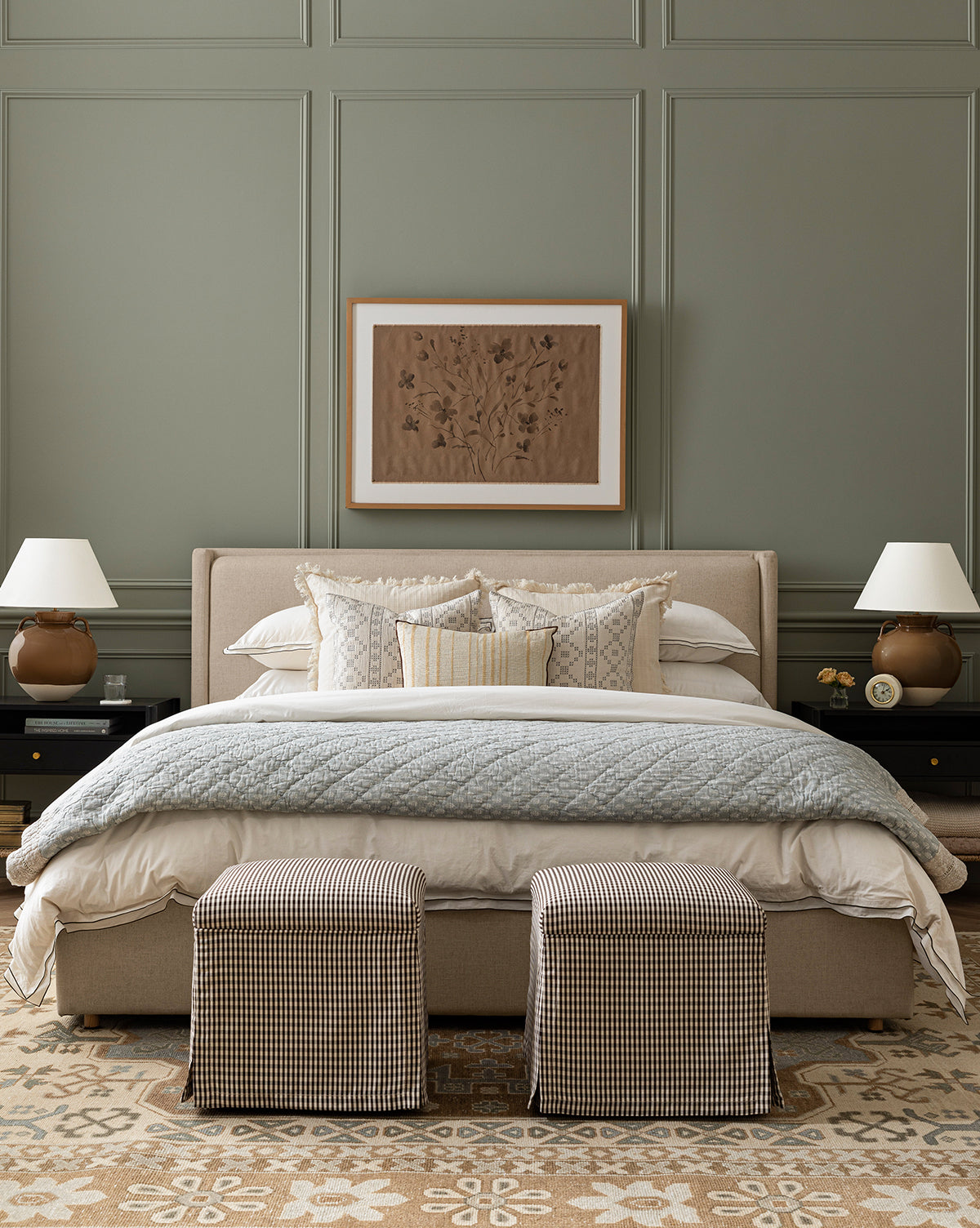 A stylish bedroom with a beige Ria Bed Low Headboard, layered white and patterned pillows, a quilted blanket, two checkered stools at the foot, matching bedside tables with lamps, and framed artwork on a green-paneled wall by McGee & Co.